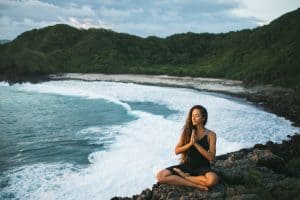 Young woman praying and meditating alone at sunset with beautiful ocean and mountain view